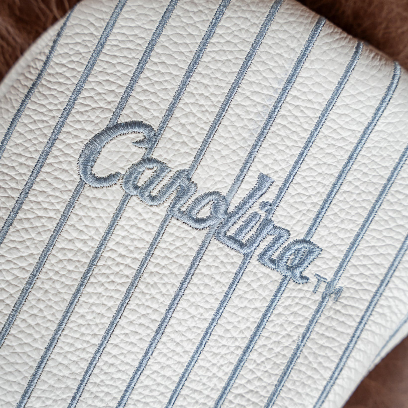 Close-up of a white textured leather item with thin blue pinstripes and an embroidered blue 'Carolina' script logo.